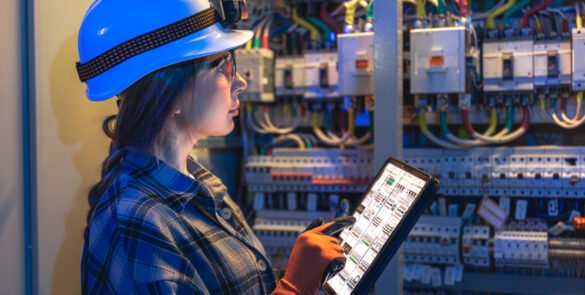 A woman electrician in a hardhat and gloves inspects power modules and wiring inside an open cabinet. Blue and warm light support smart monitoring, industry 4.0 and safety compliance.